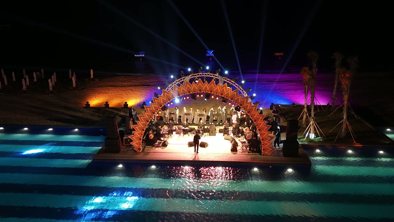 An aerial view of a waterfront evening at King Abdullah Economic City: a live stage under orange geometric arches, a pool striped with turquoise light, and guests gathered along the water.