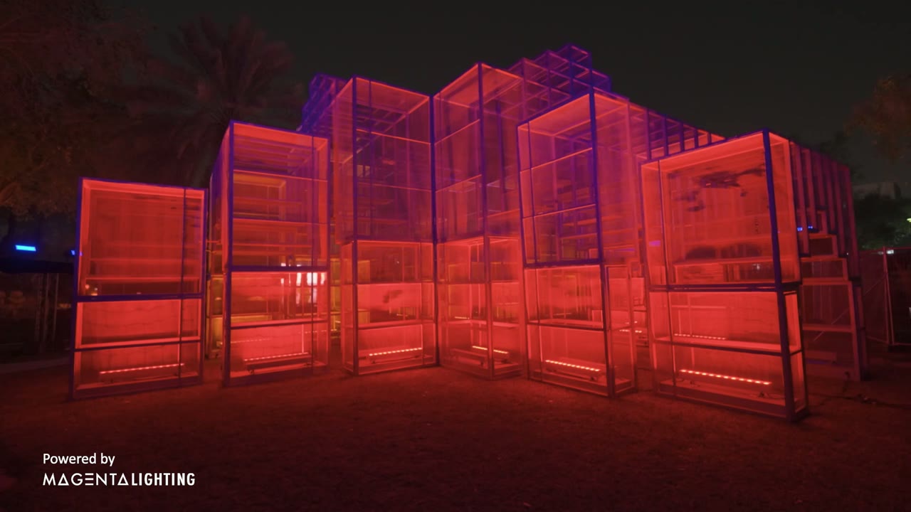 A walk-around sculpture in Riyadh: stacked steel-and-mesh volumes lit amber from within, standing on grass at night.