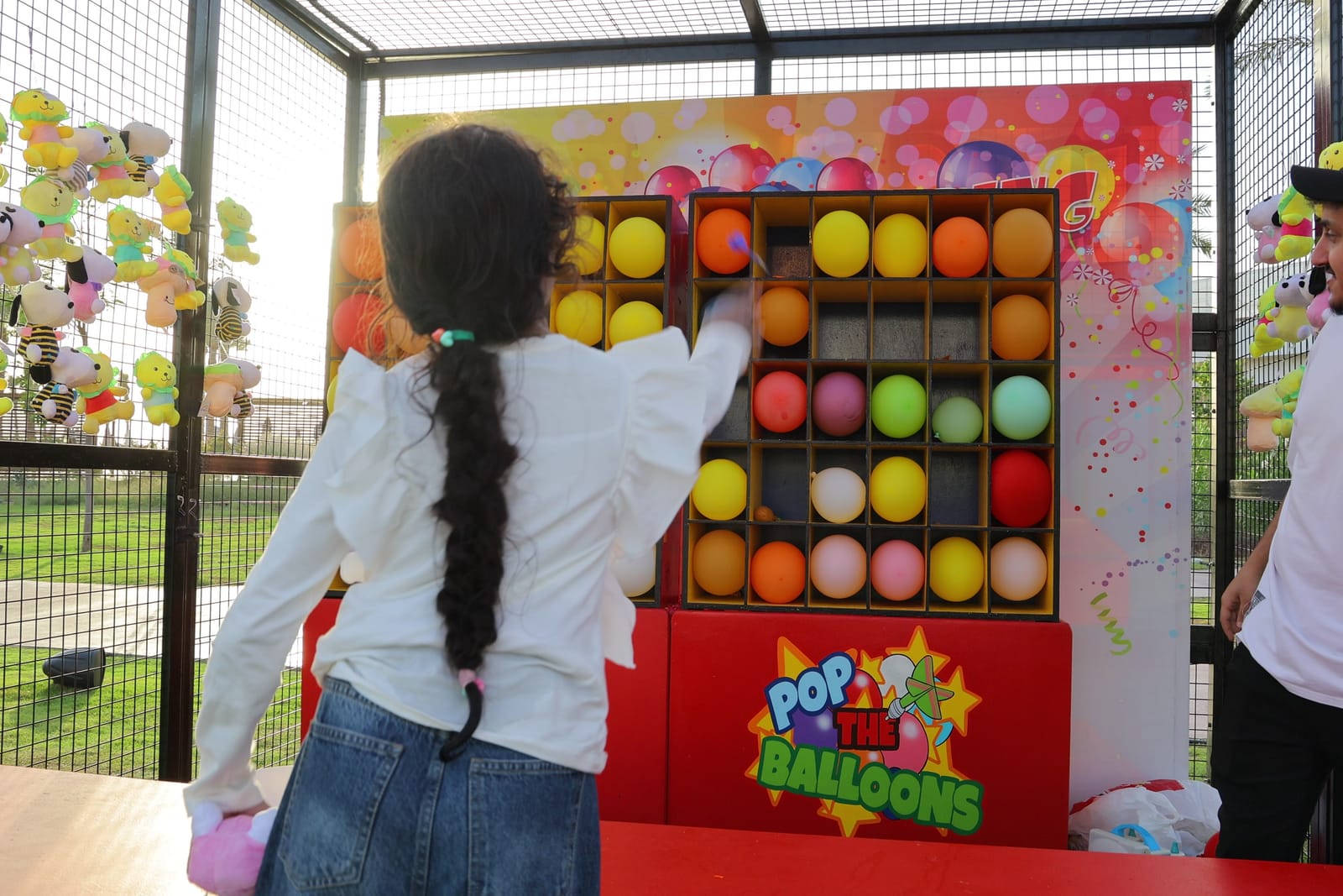 A girl throwing at a wall of coloured balloons at a carnival game stall.