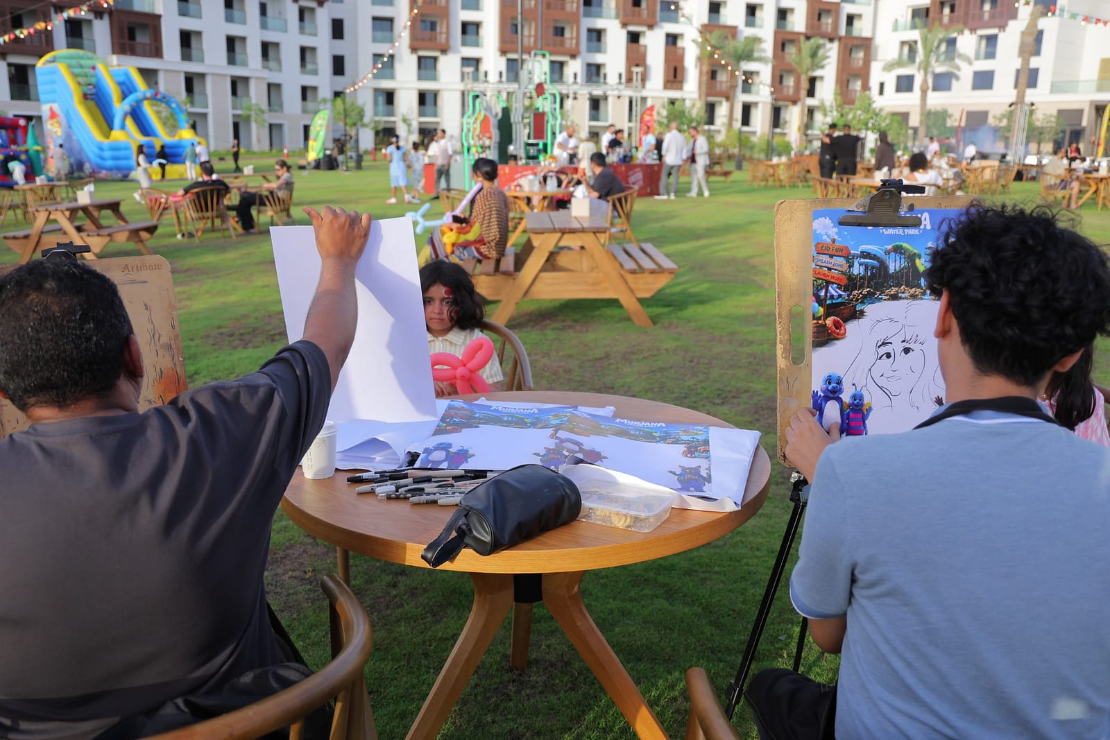 Two caricature artists drawing guests at a table on the carnival lawn.
