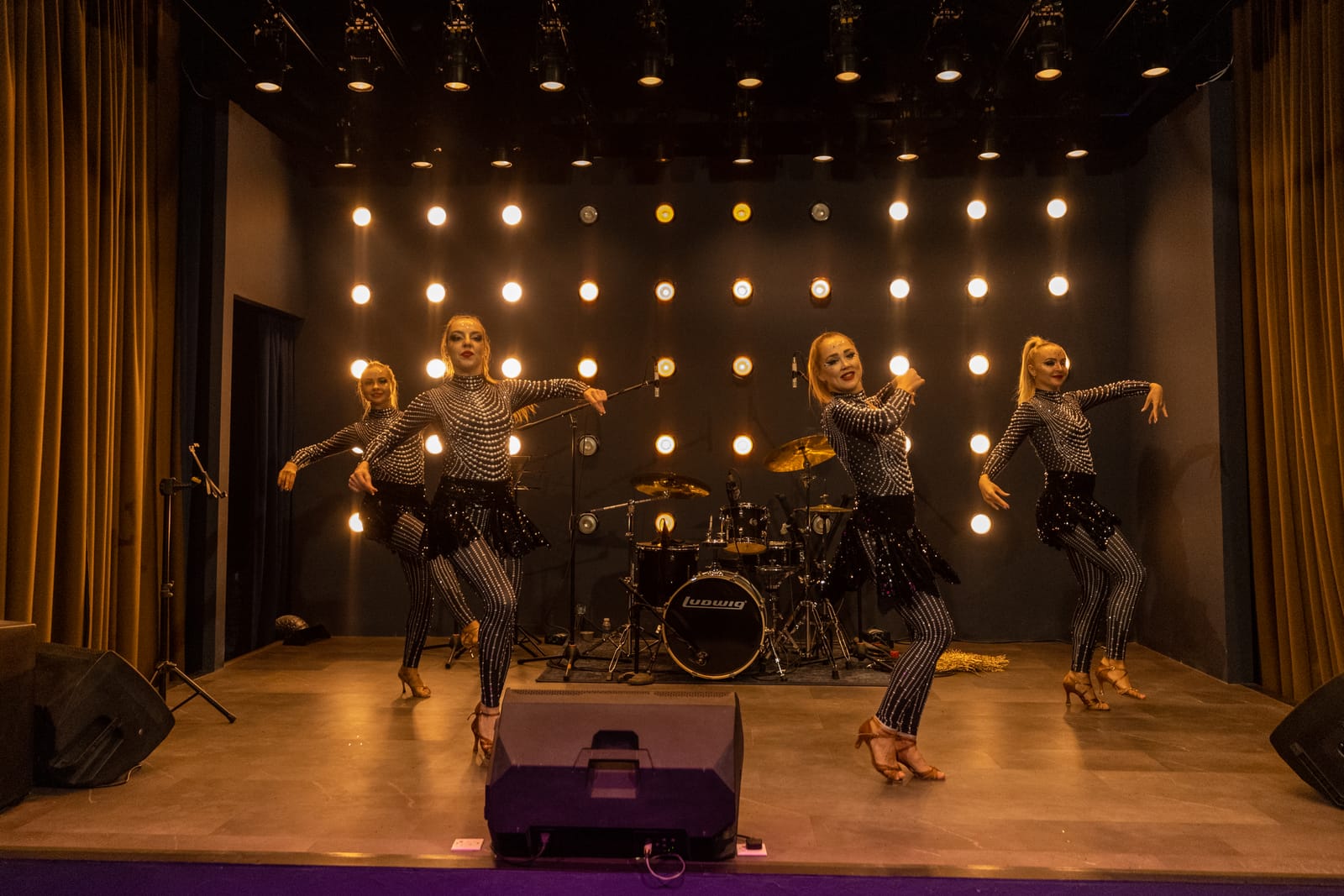 Four dancers mid-routine on Zodiac Stage, in front of a wall of warm stage lights and a drum kit.