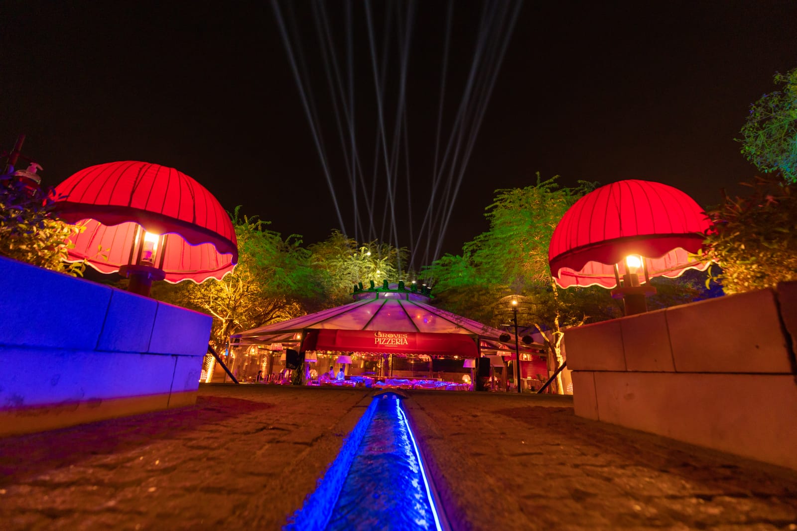 A garden pavilion at night between red canopies, a lit water channel leading to it and beams rising behind.