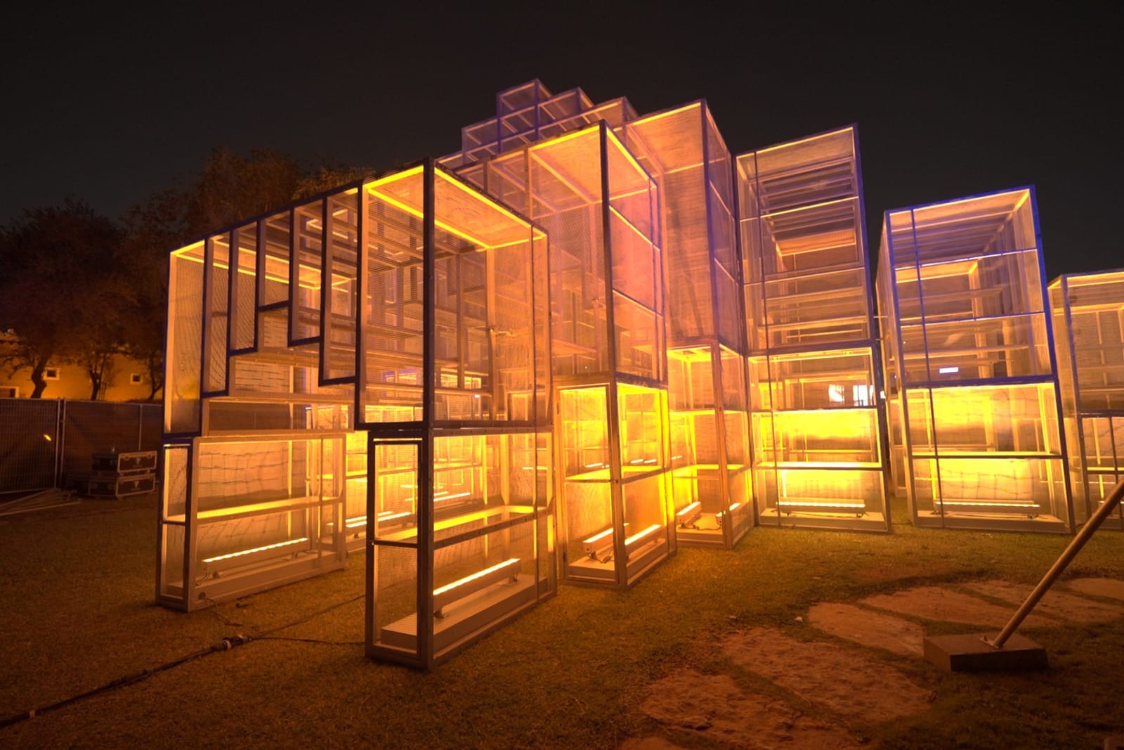 A sculpture of stacked steel and mesh volumes lit amber from within, on grass at night.