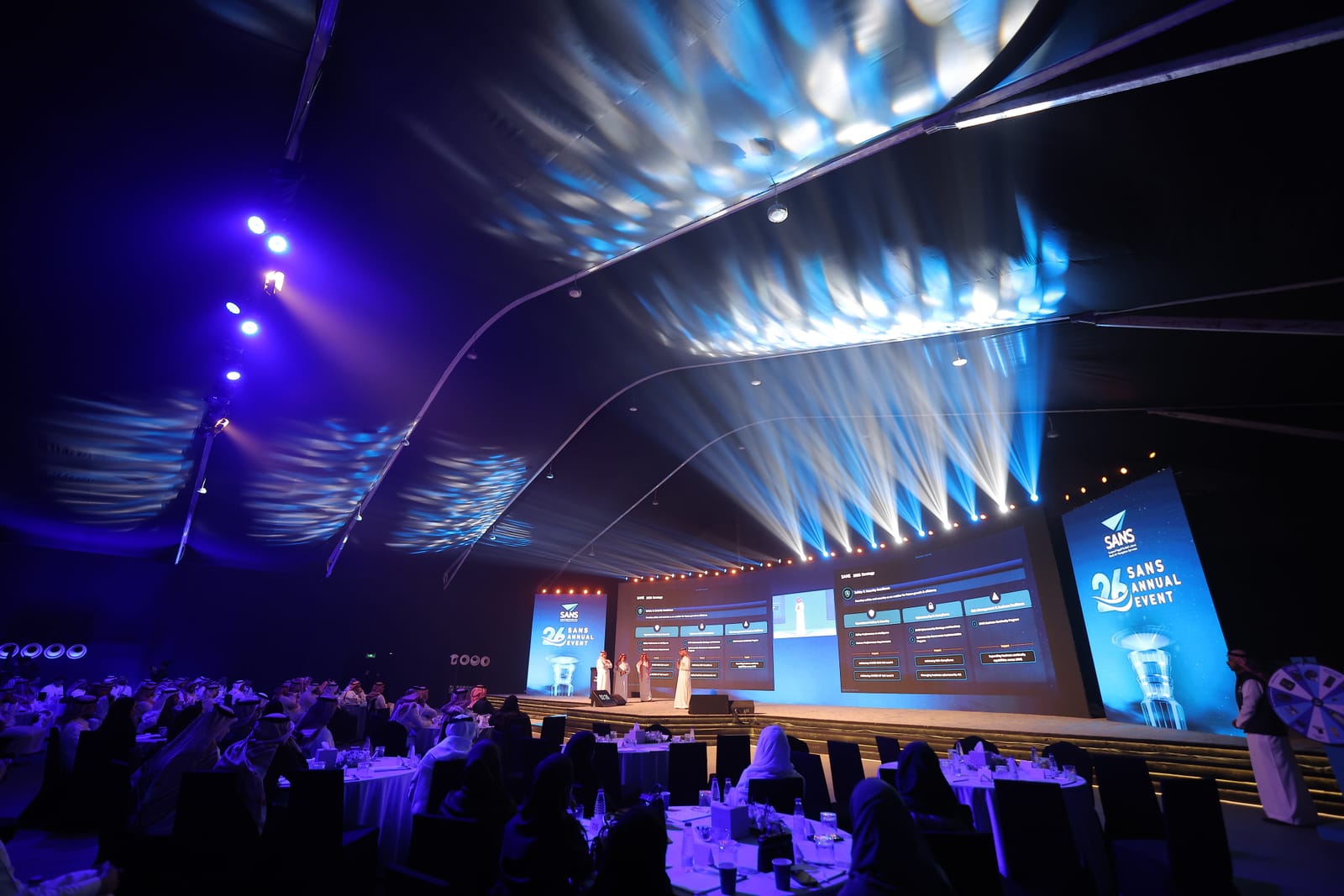 A large event hall at night: guests at round tables facing a wide stage with three screens under blue beams.