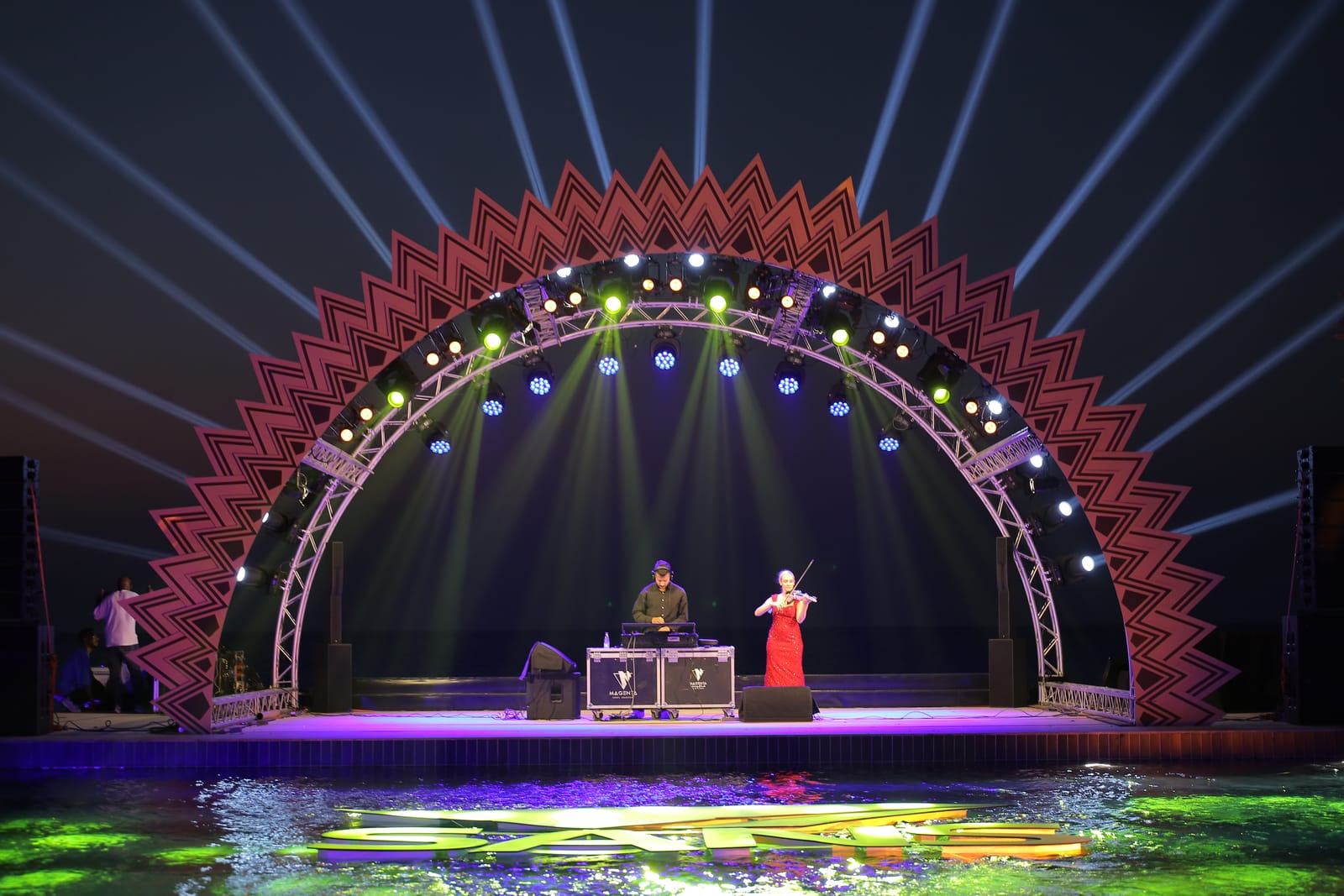 An arched stage over water at night with beams fanning out behind it and two performers on stage.