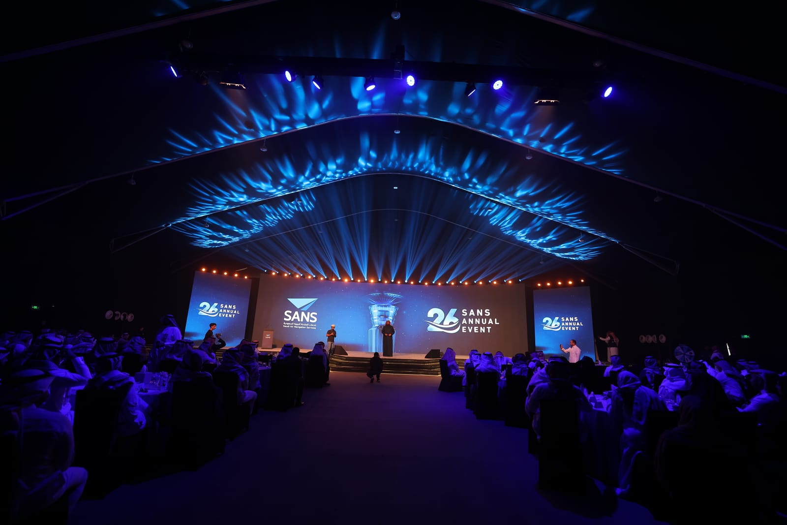 A hall seen from the back: an audience facing a stage with screens under layered blue beams.