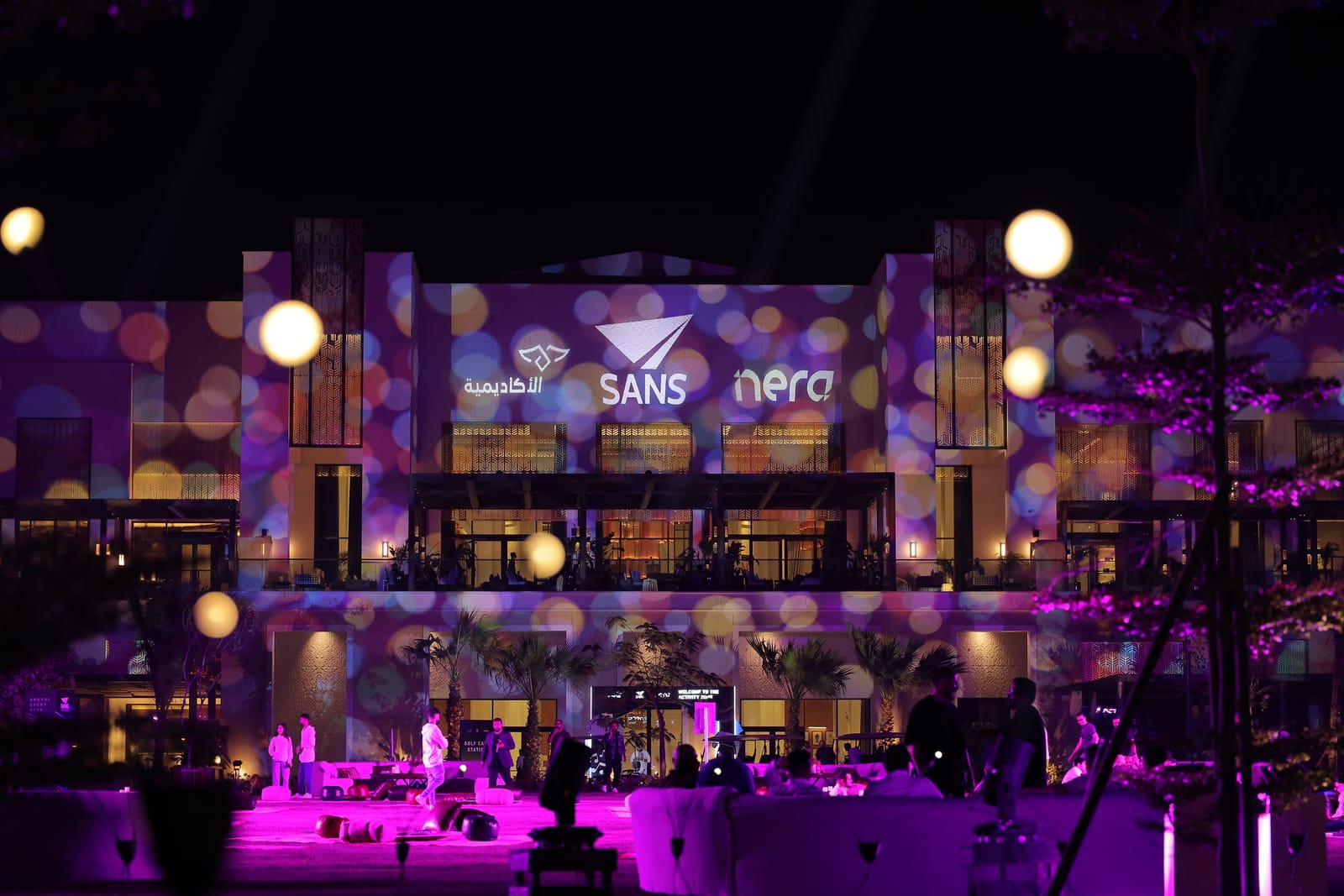 A building front covered in projected light and logos at night, with guests on the terrace below.