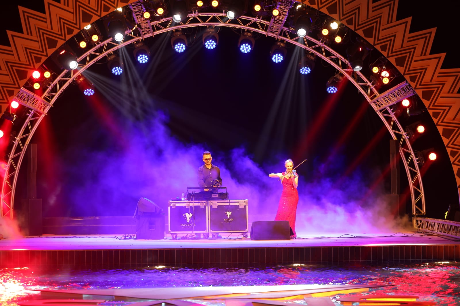 A DJ and a violinist on an arched stage, lit red and violet through low smoke over the water.