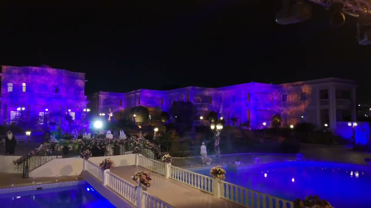 A beach house at night seen from the water: the facade washed in colour, reflections across the sea, and a lit pool terrace in front of it.